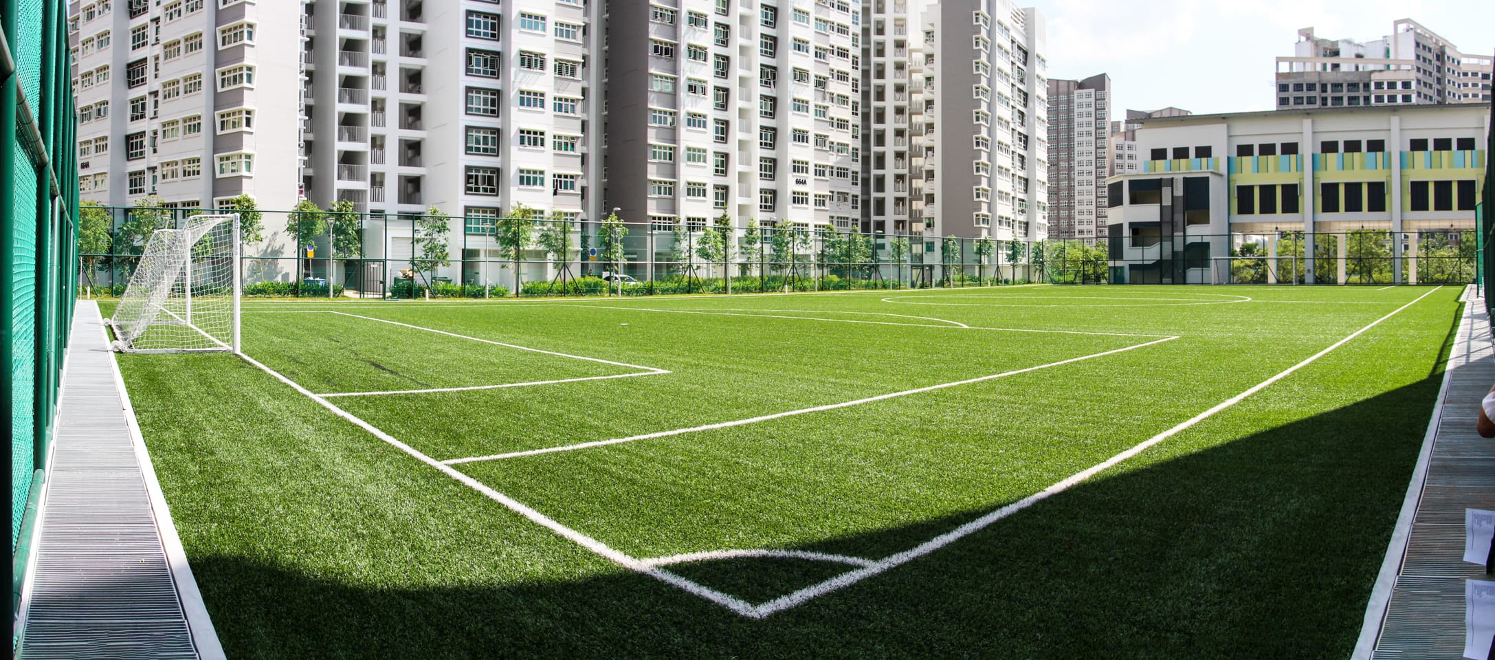 Empty outdoor soccer field with artificial turf and a white goal, surrounded by tall residential apartment buildings.